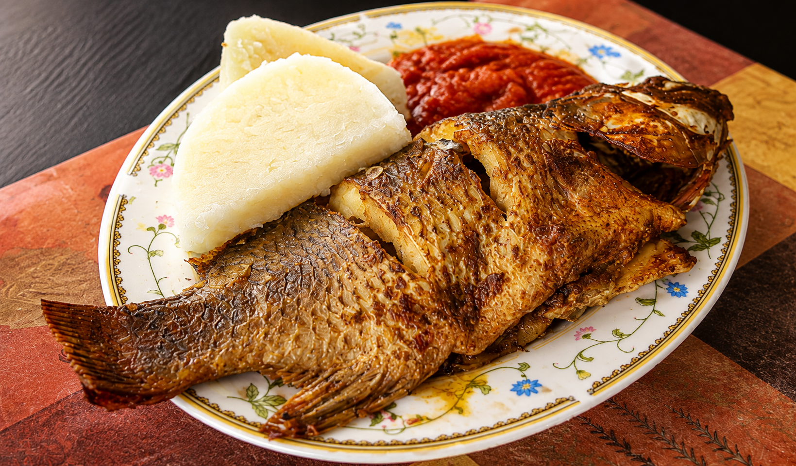 Whole fried fish with boiled yam wedges and red pepper stew on a patterned plate
